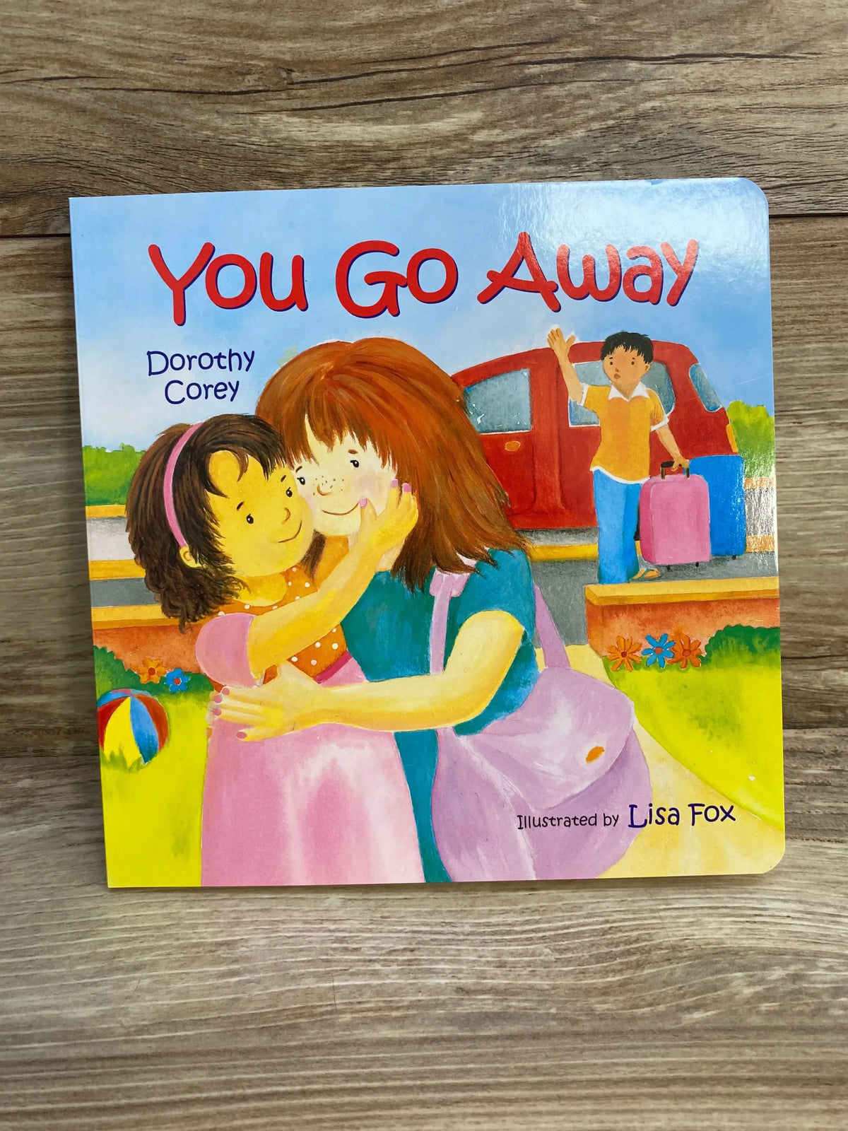 Front cover of You Go Away board book showing two girls hugging with a boy waving by a red car on a wooden surface