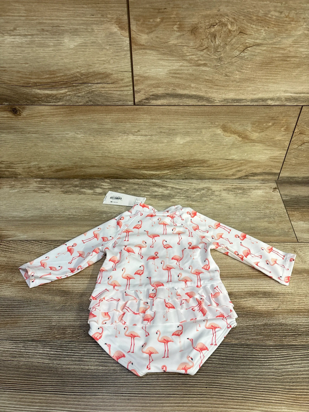 Flat lay of a white Janie and Jack flamingo rash guard swimsuit showcasing long sleeves and ruffled bottom on wood