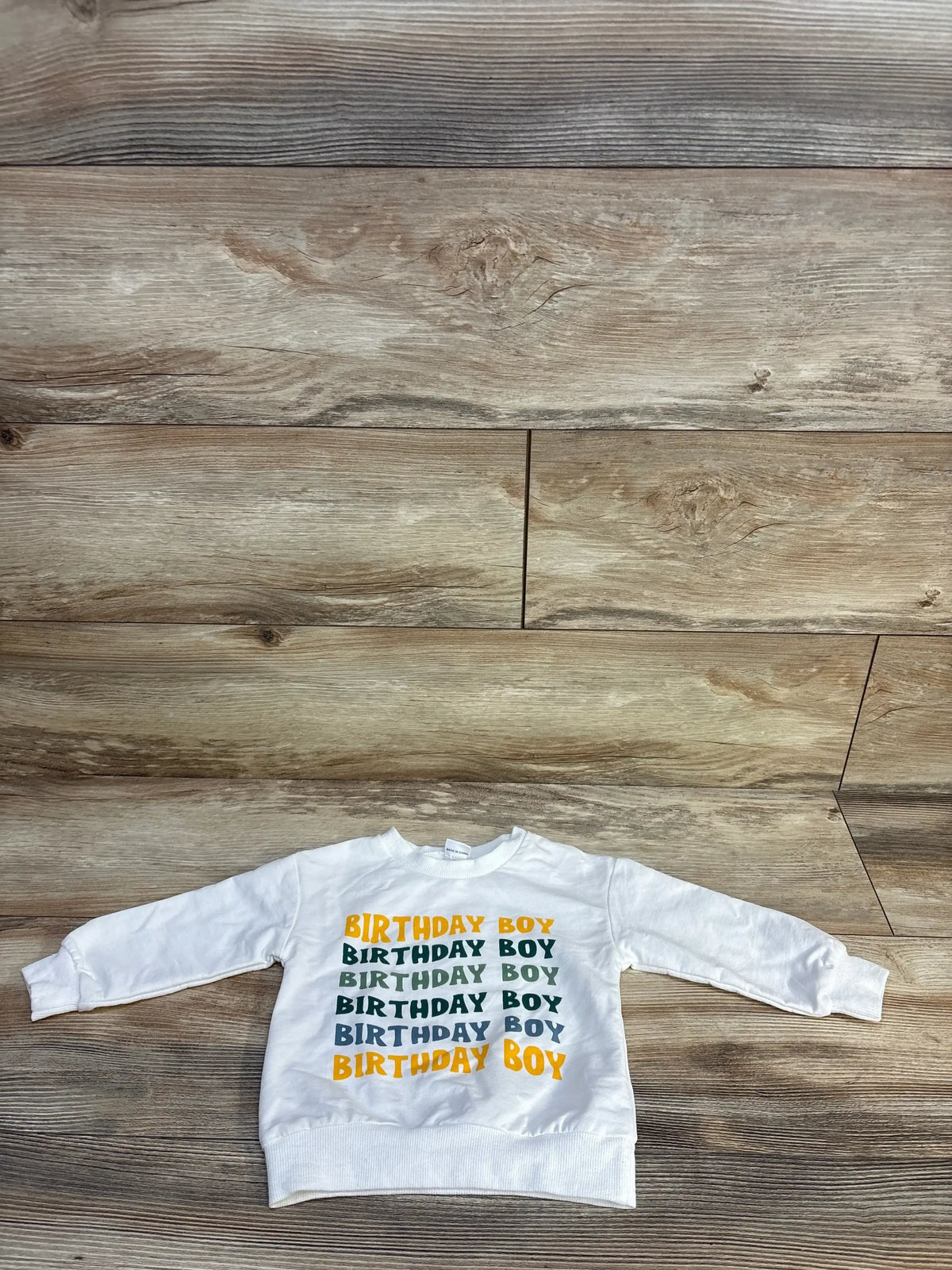 White long-sleeve kids sweatshirt with 'Birthday Boy' text in green and yellow on wood floor