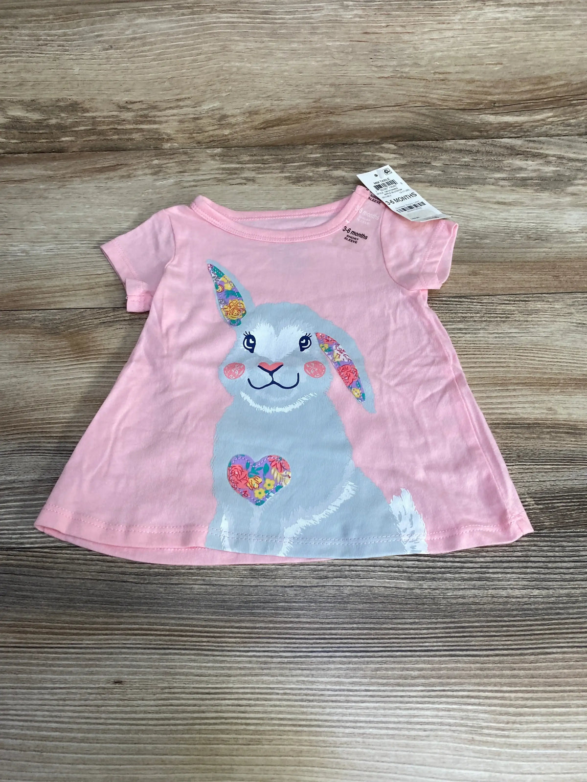Flat-lay of pink baby shirt (3-6 months) showing large gray bunny graphic with floral ears and heart on wood background
