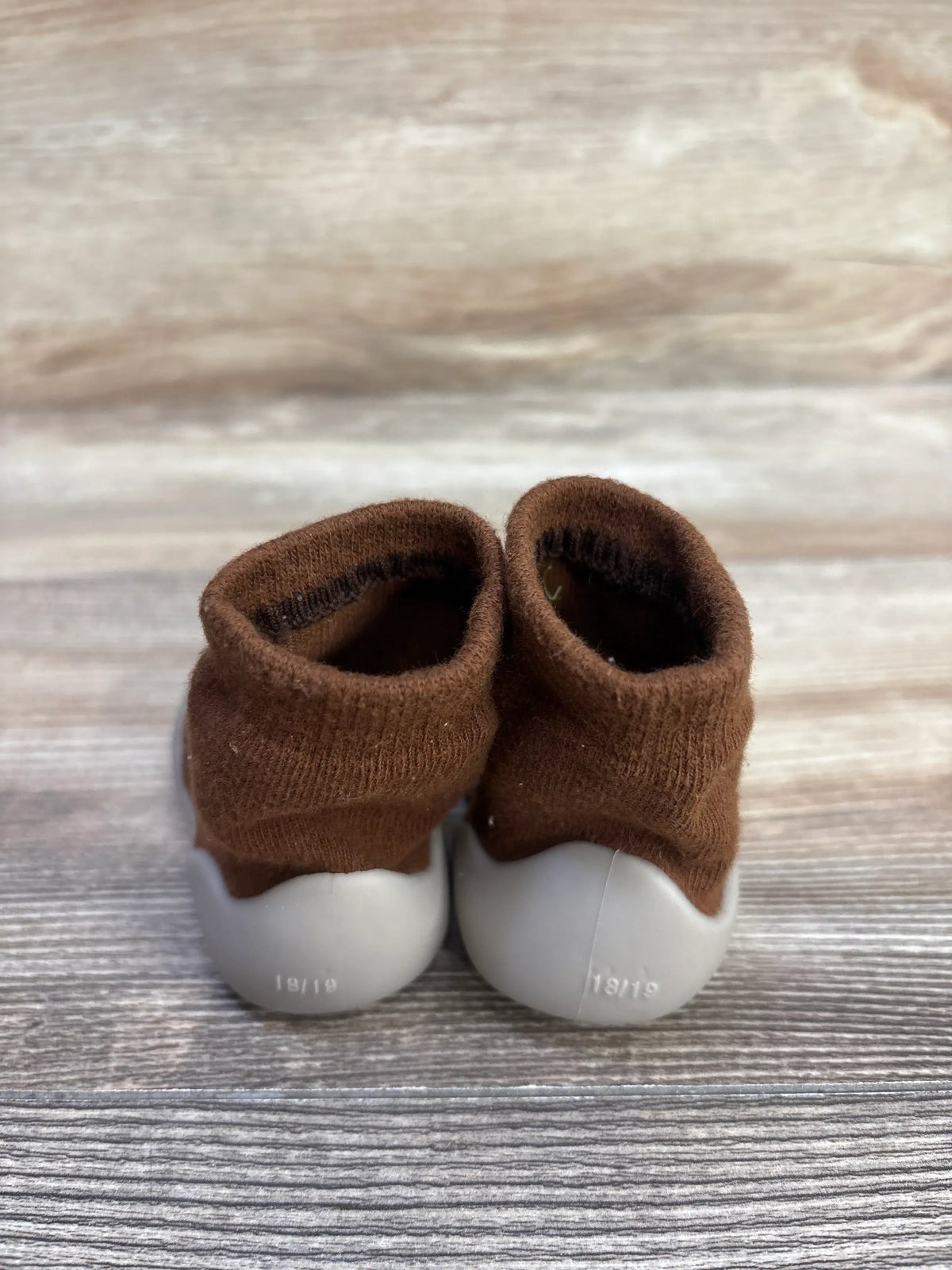 Rear view of brown Tiny Explorings self-care sock shoes showing gray soles marked 18/19 on a wood-grain surface