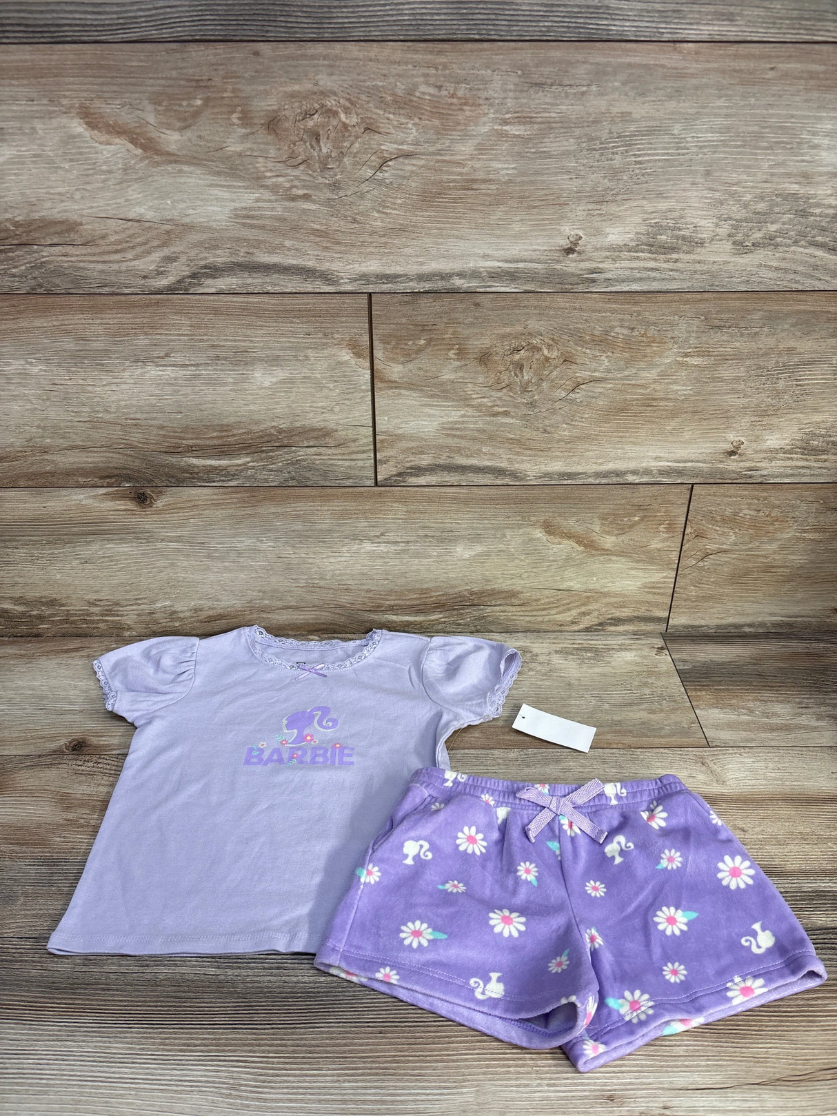 Flat lay of lilac Barbie 2-piece tee and shorts showing logo on tee, lace trim, and floral shorts with bow on wood background