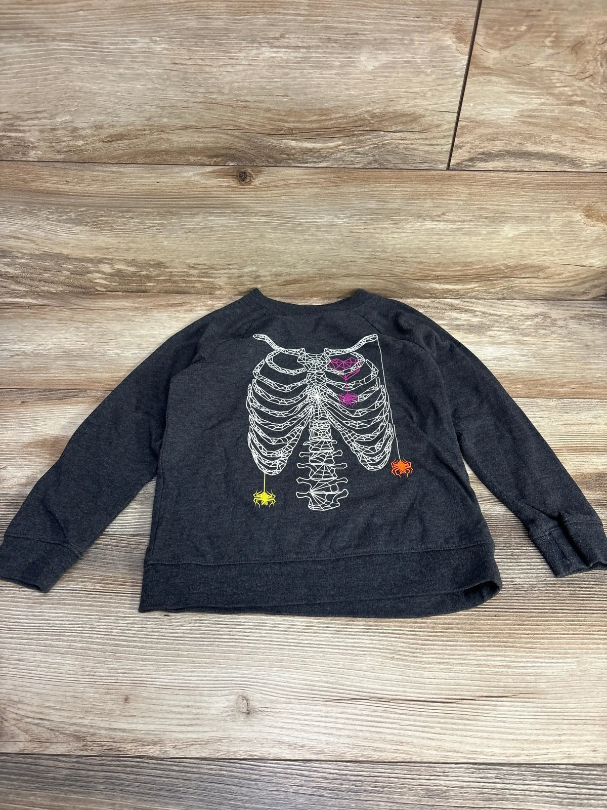 Flat lay of Cat & Jack grey long-sleeve toddler skeleton shirt showing white ribcage graphic and three small colored spiders on wood floor background