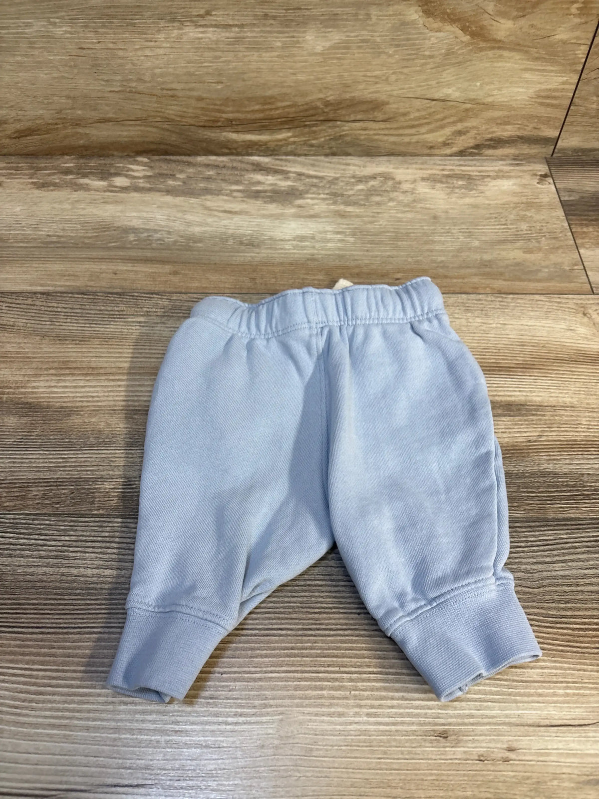 Top-down view of light blue baby jogger pants (3M) showing elastic waistband and ribbed cuffs on wood floor background