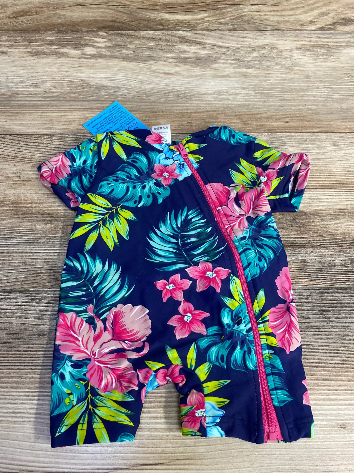 Flat lay of navy baby one-piece swimsuit showing tropical floral print and pink diagonal front zipper, size 3M tag on wood background