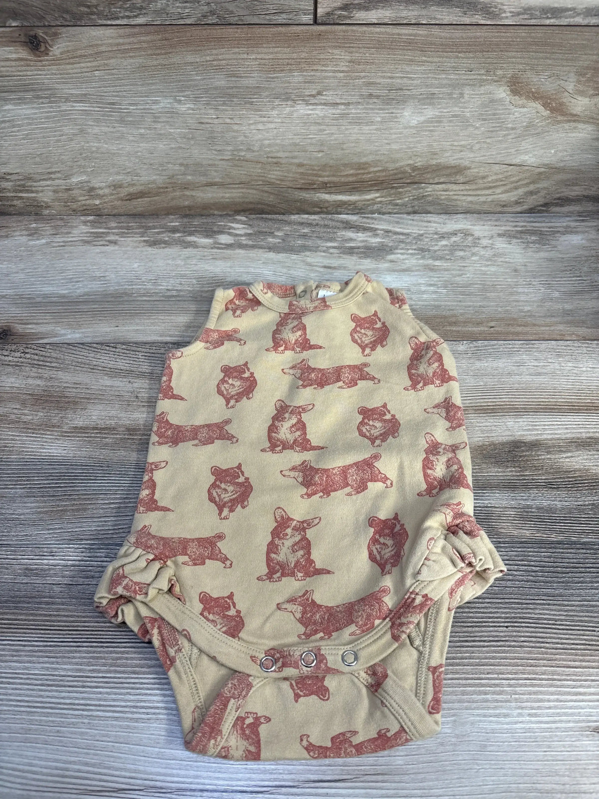 Flat lay of tan Kate Quinn Korgi bodysuit with red corgi print, ruffle leg openings and three-snap crotch on wood background