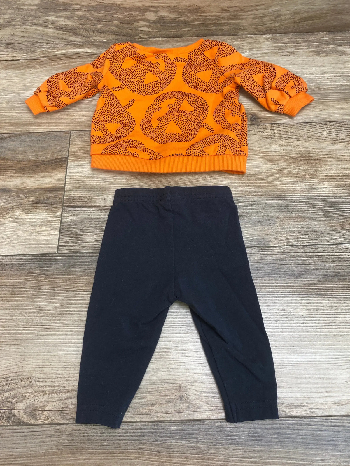 Front view of Carter’s 2-piece size 3M outfit showcasing an orange jack-o-lantern shirt and black pants on wood flooring