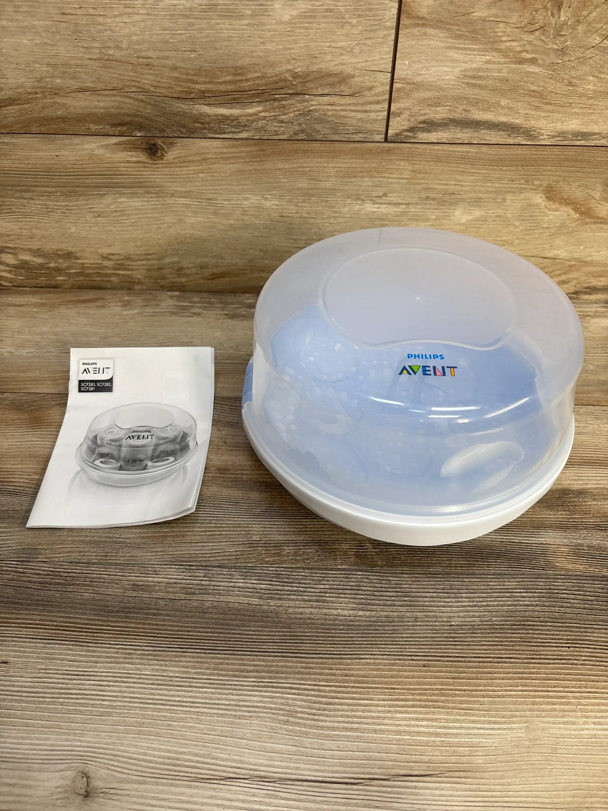 Three-quarter view of AVENT microwave sterilizer with translucent lid and white base, instruction booklet on wood surface
