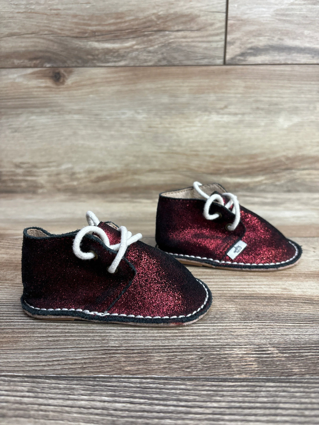 Pair of small toddler shoes with burgundy glitter fabric and white laces on a wooden floor