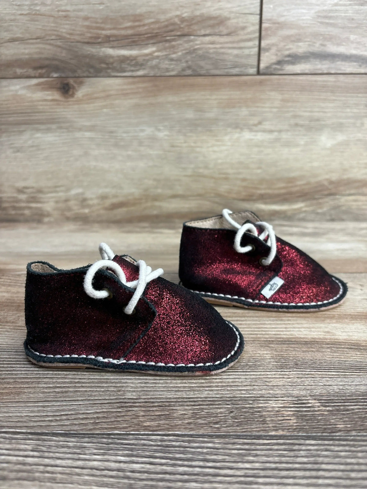 Pair of small toddler shoes with burgundy glitter fabric and white laces on a wooden floor