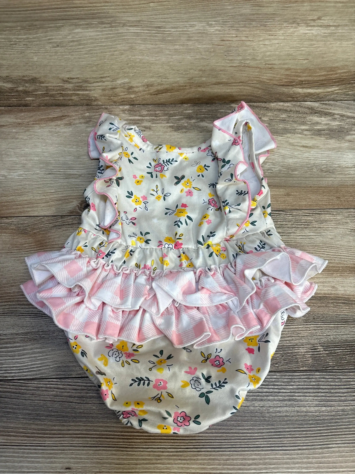 Top-down view of ivory floral bubble romper showcasing pink gingham ruffle tiers and flutter sleeves on wood background