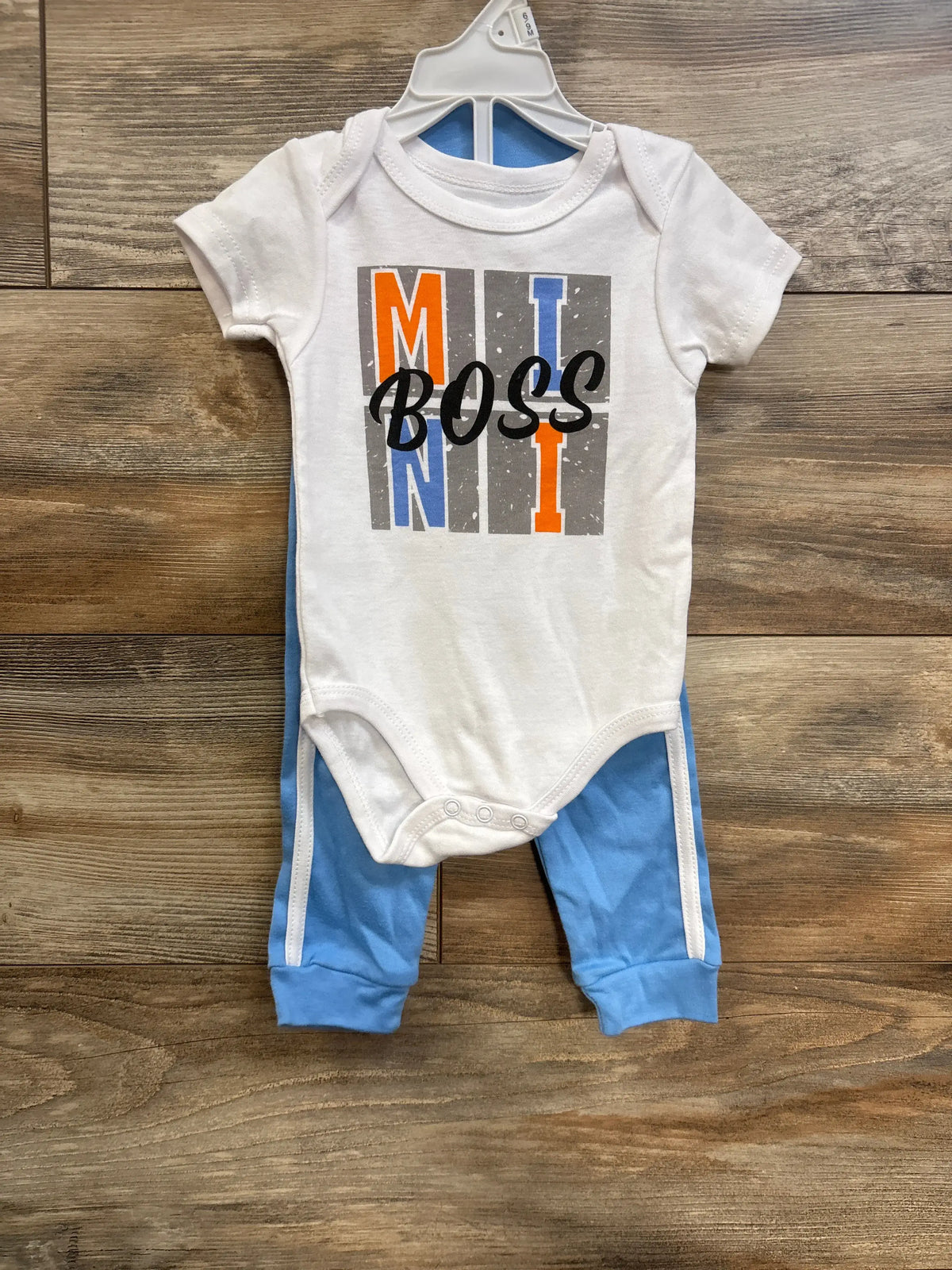 Front view of white baby bodysuit and blue pants showcasing BOSS graphic on bodysuit with wood floor background