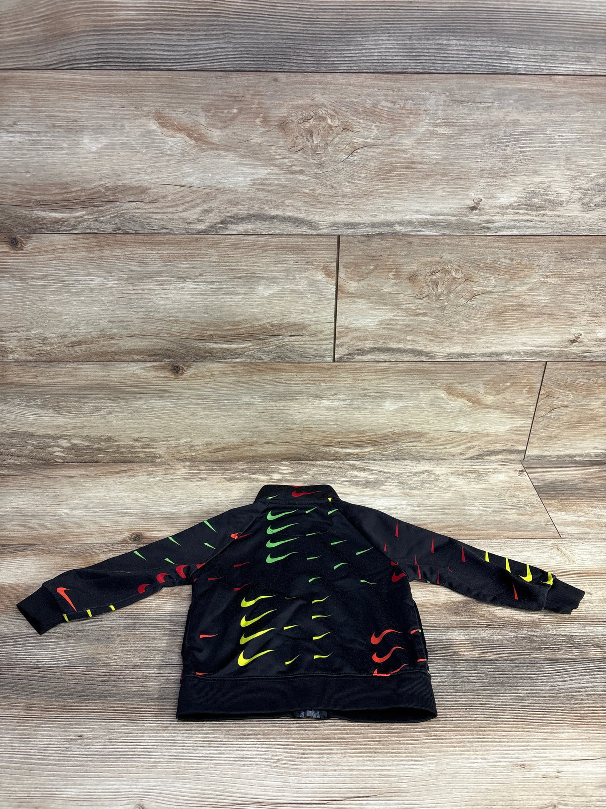 Black kids Nike jacket with colorful red, yellow, and green swoosh logos laid flat on wooden floor