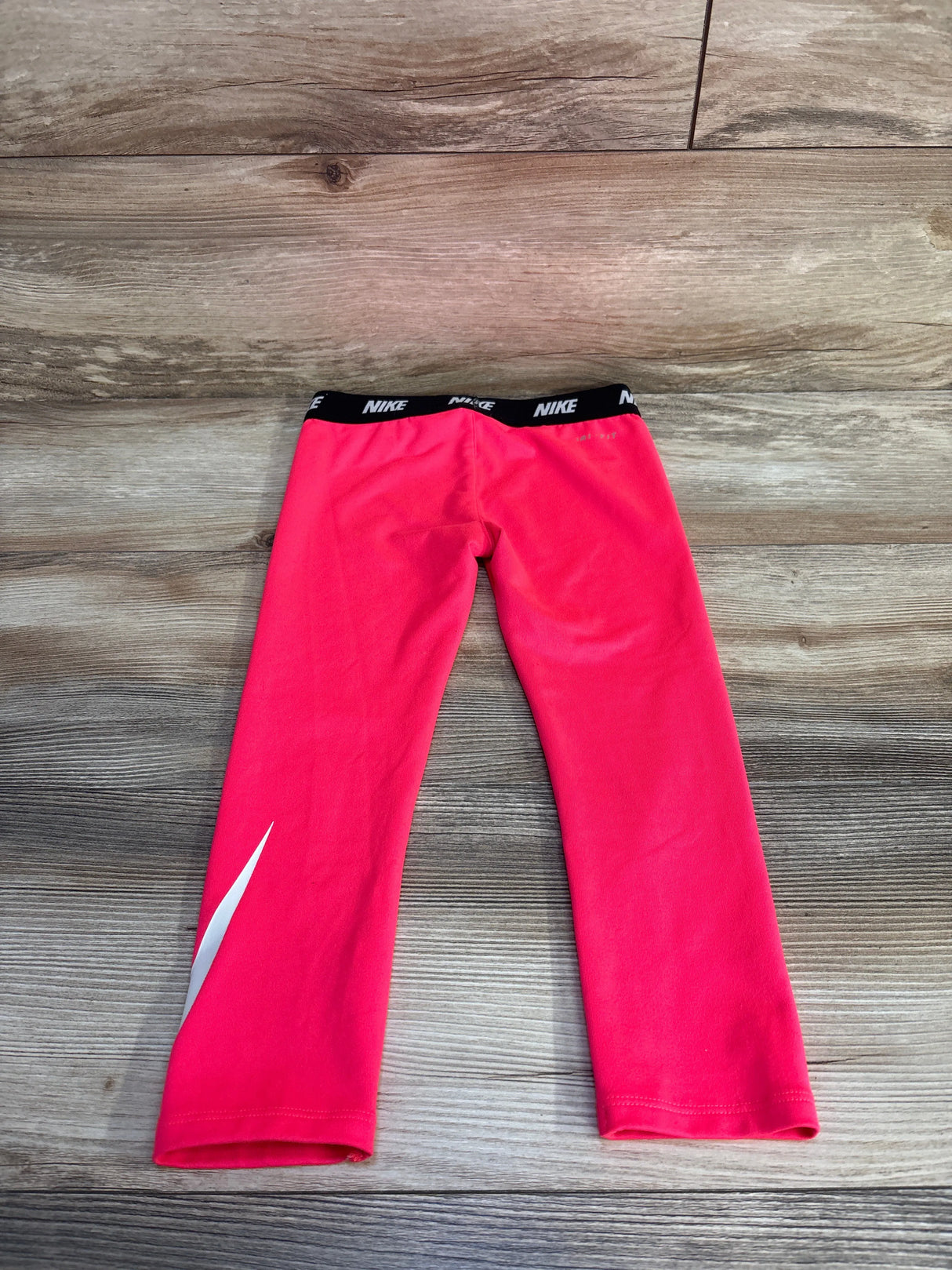 Flat lay back view of girls Nike Dri-Fit leggings size 4T showing black elastic waistband with repeating Nike logo and white swoosh on wood floor