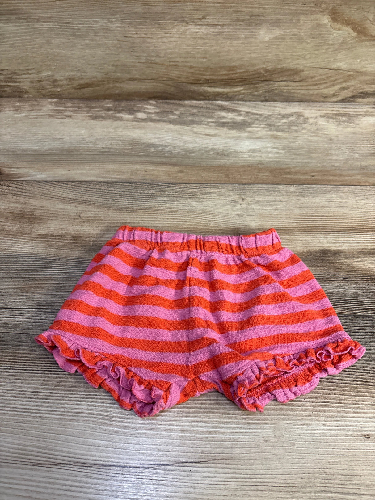 Flat lay of toddler pink and orange striped shorts showing elastic waistband and ruffled hems on wood background