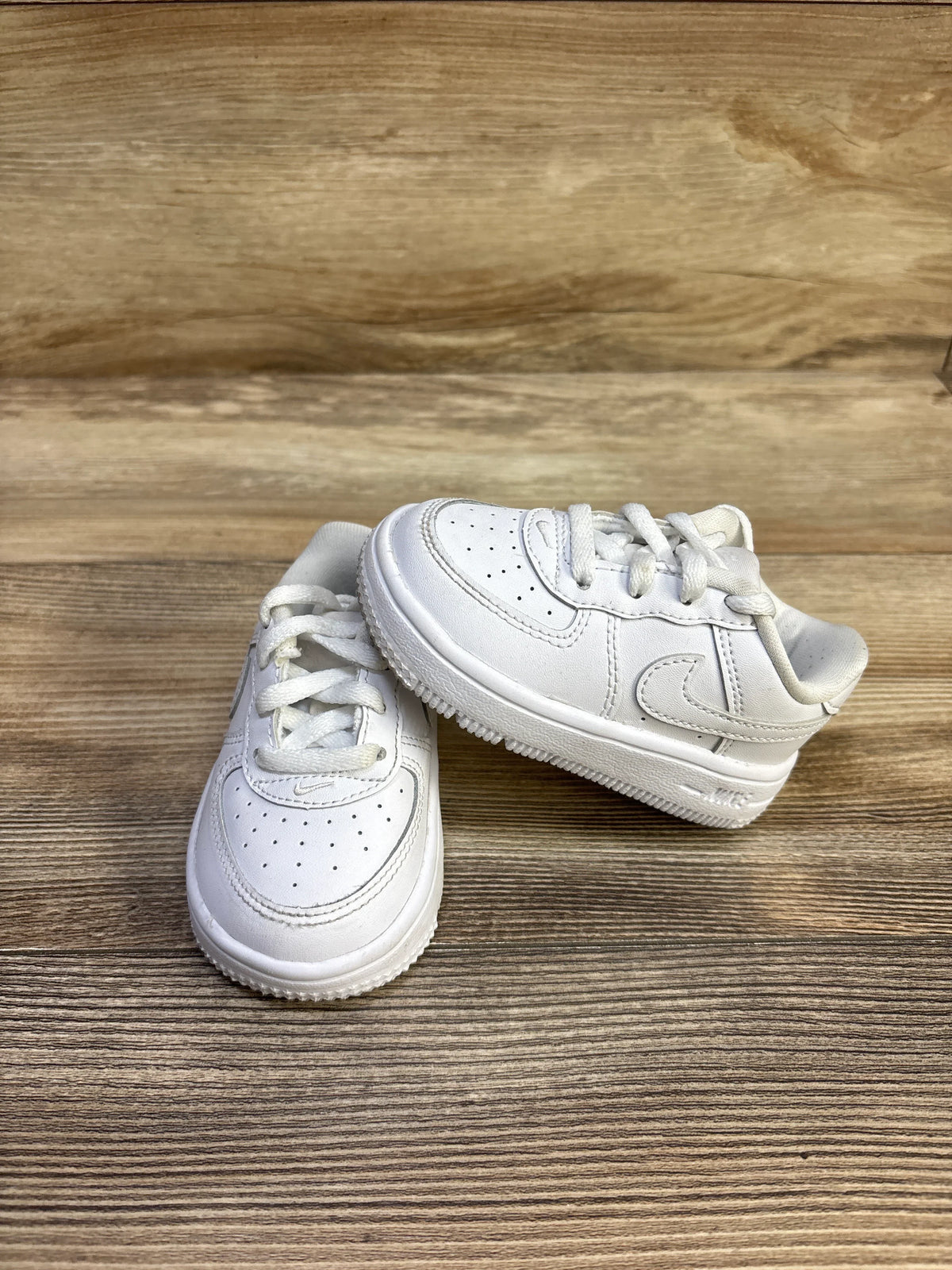 Front and side view of toddler white Nike Air Force 1 Low sneakers showcasing perforated toe, stitched Swoosh, and laces on light wood background