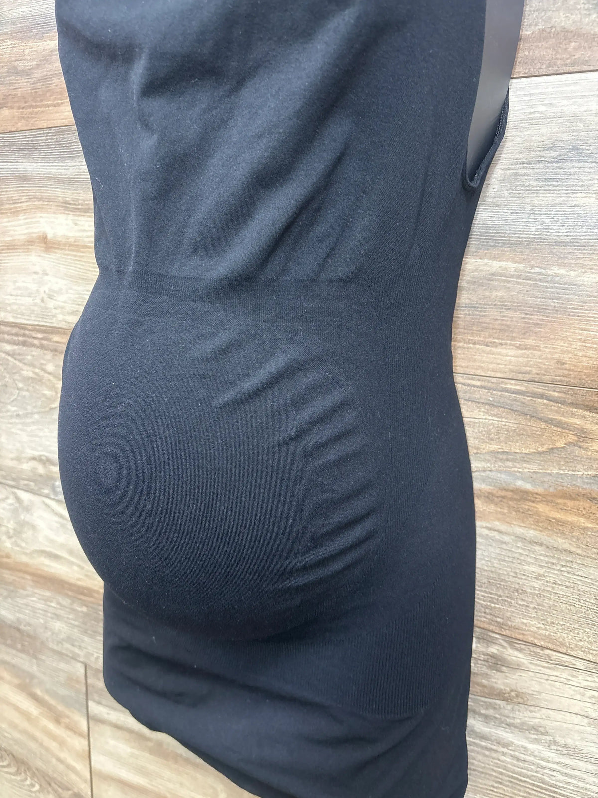 Close-up front view of black Blanqi maternity belly support tank top showcasing its stretchy belly panel on a wood floor
