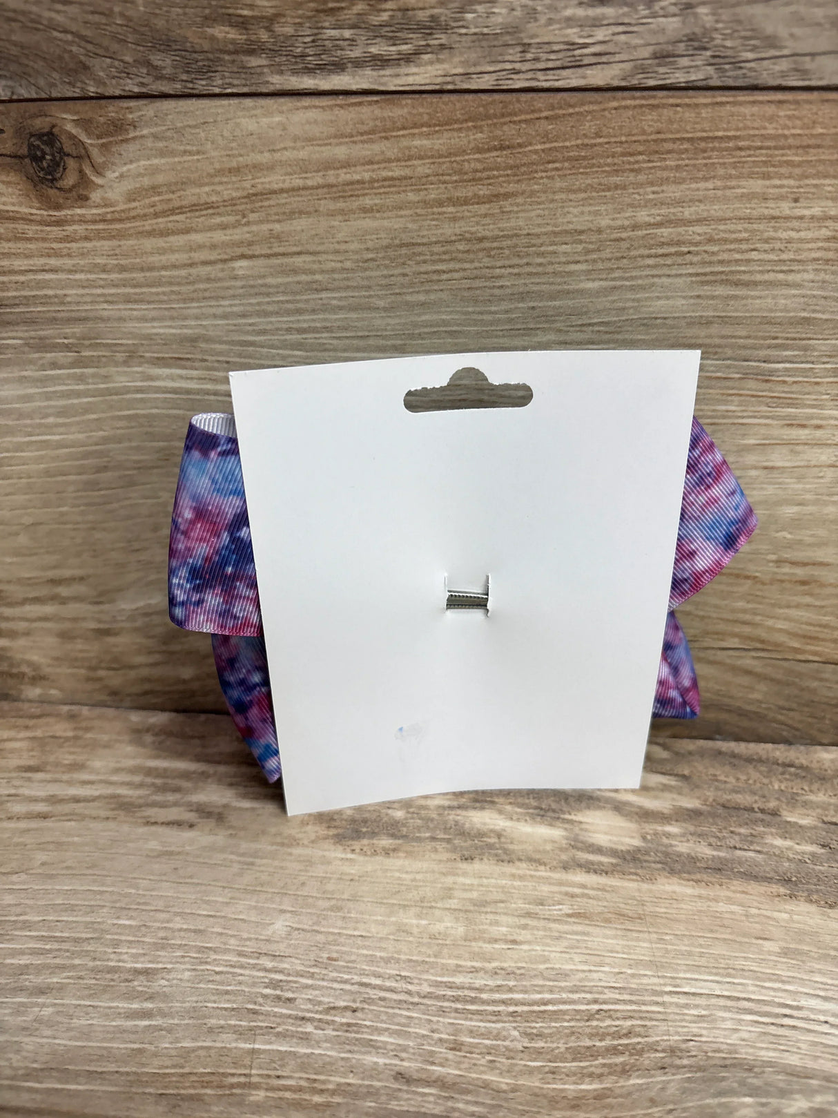 Rear view of large galaxy purple hair bow with tie-dye pattern and silver alligator clip on white card and wood background