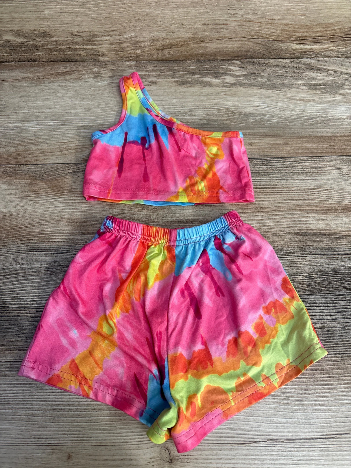 Flat lay of multicolor tie-dye baby two-piece: one-shoulder tank and elastic shorts, size 6-9 months on wood