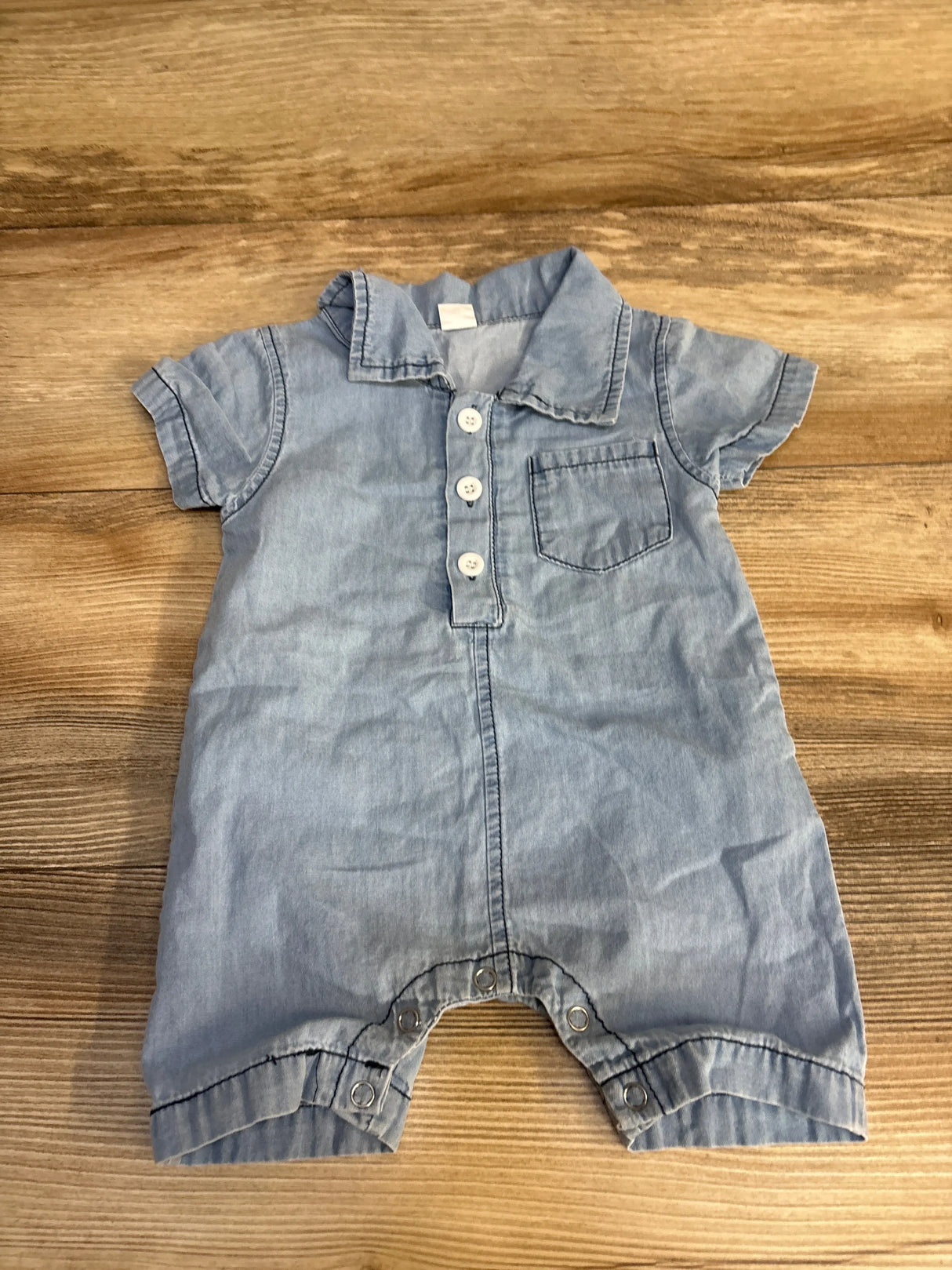 Light blue denim baby romper with short sleeves, button front, and ruffled legs on wood surface