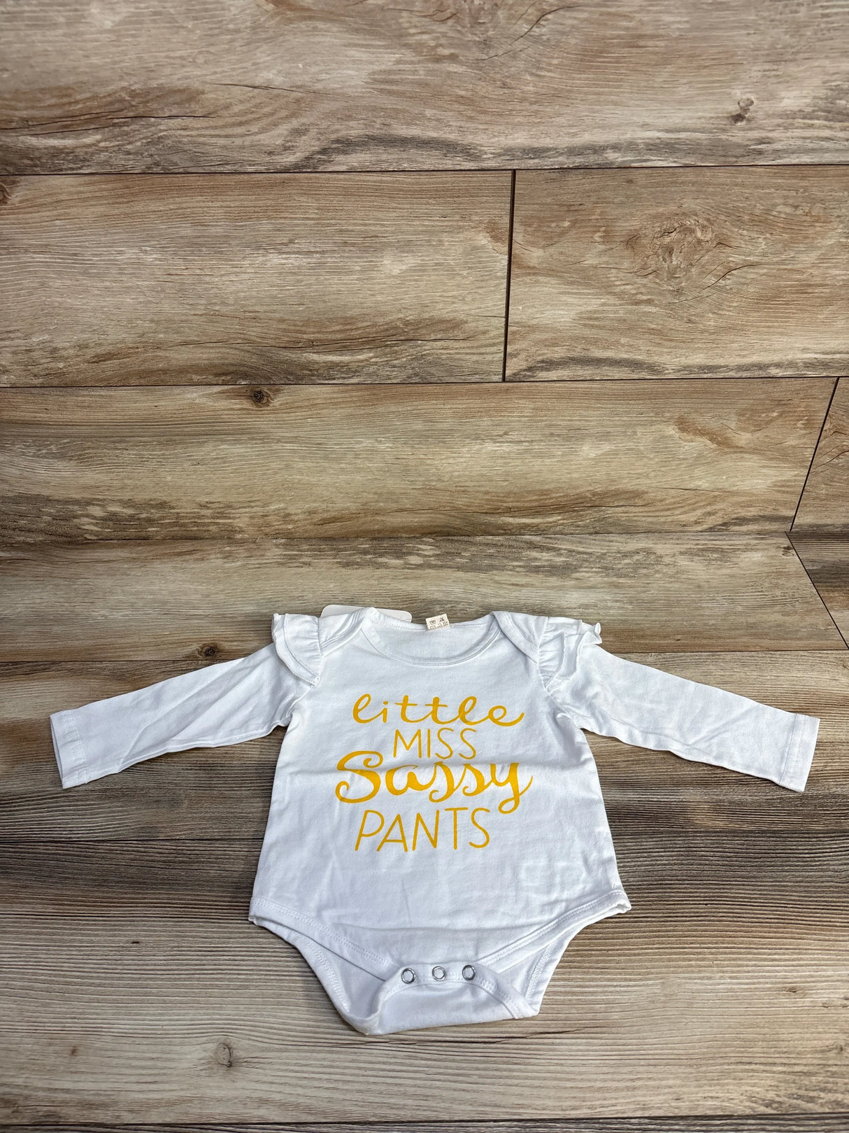White baby bodysuit with yellow 'Little Miss Sassy Pants' text on wood floor