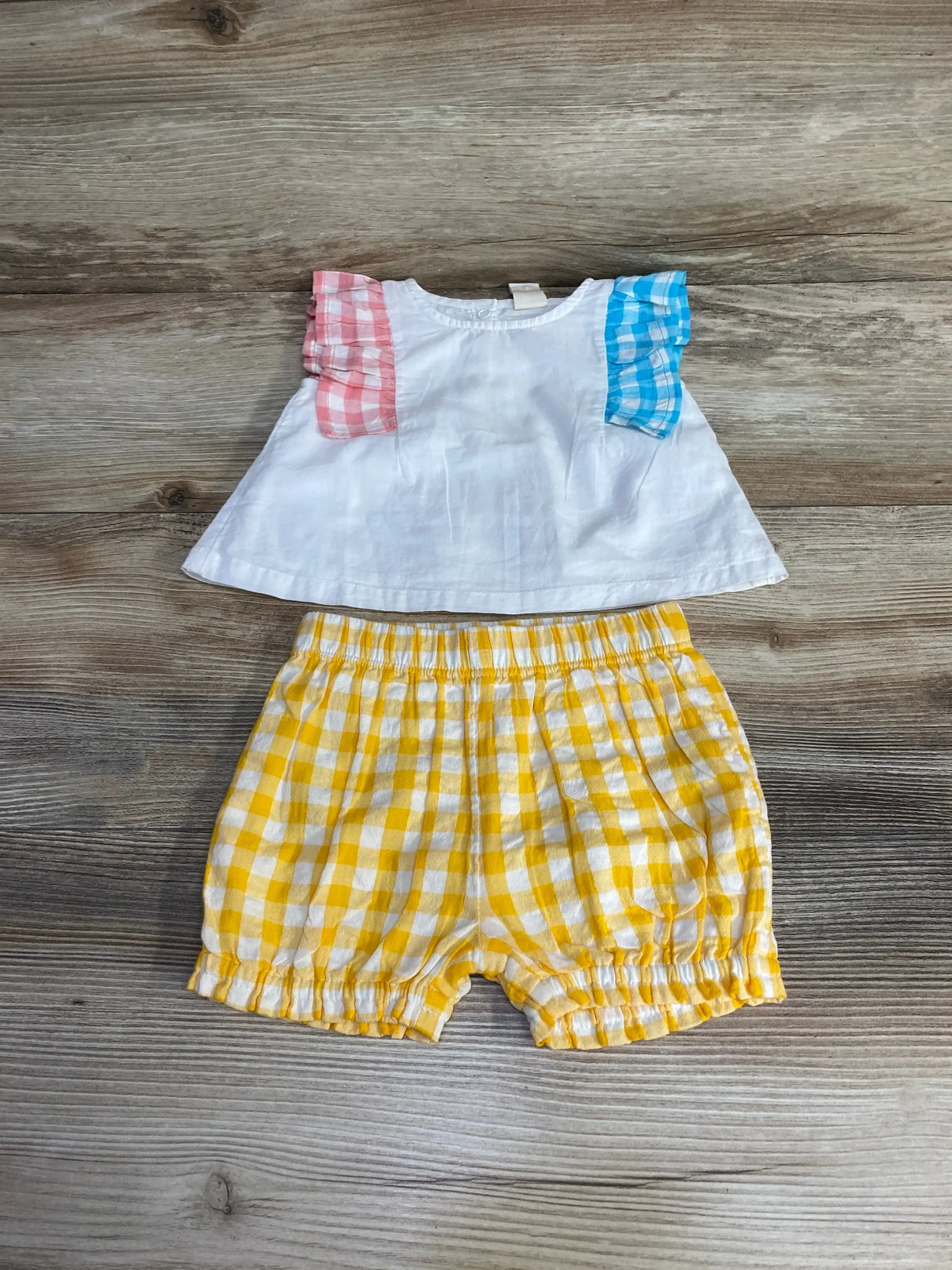 Flat lay of Tucker and Tate baby ruffle top with multicolor gingham sleeves and yellow gingham shorts on wood background