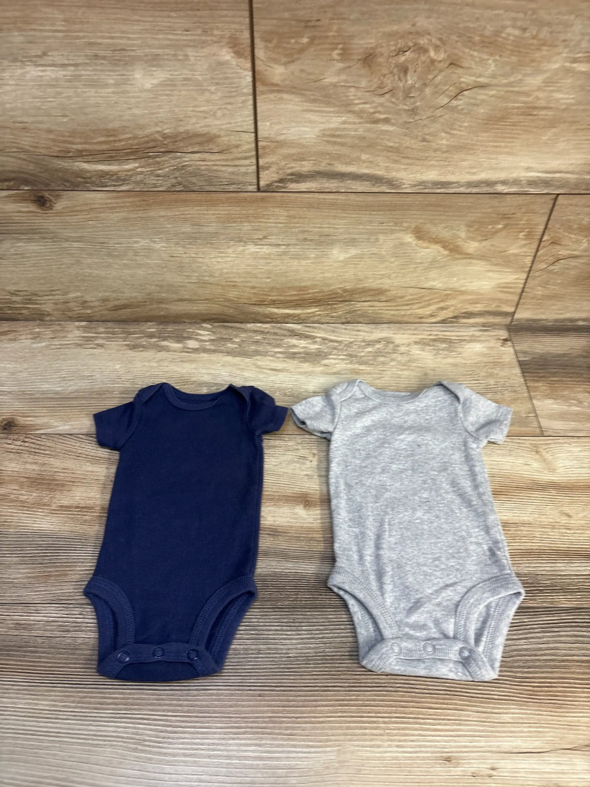 Front view of Just One You two-pack newborn bodysuits showcasing short sleeves and snap closures on a wood floor
