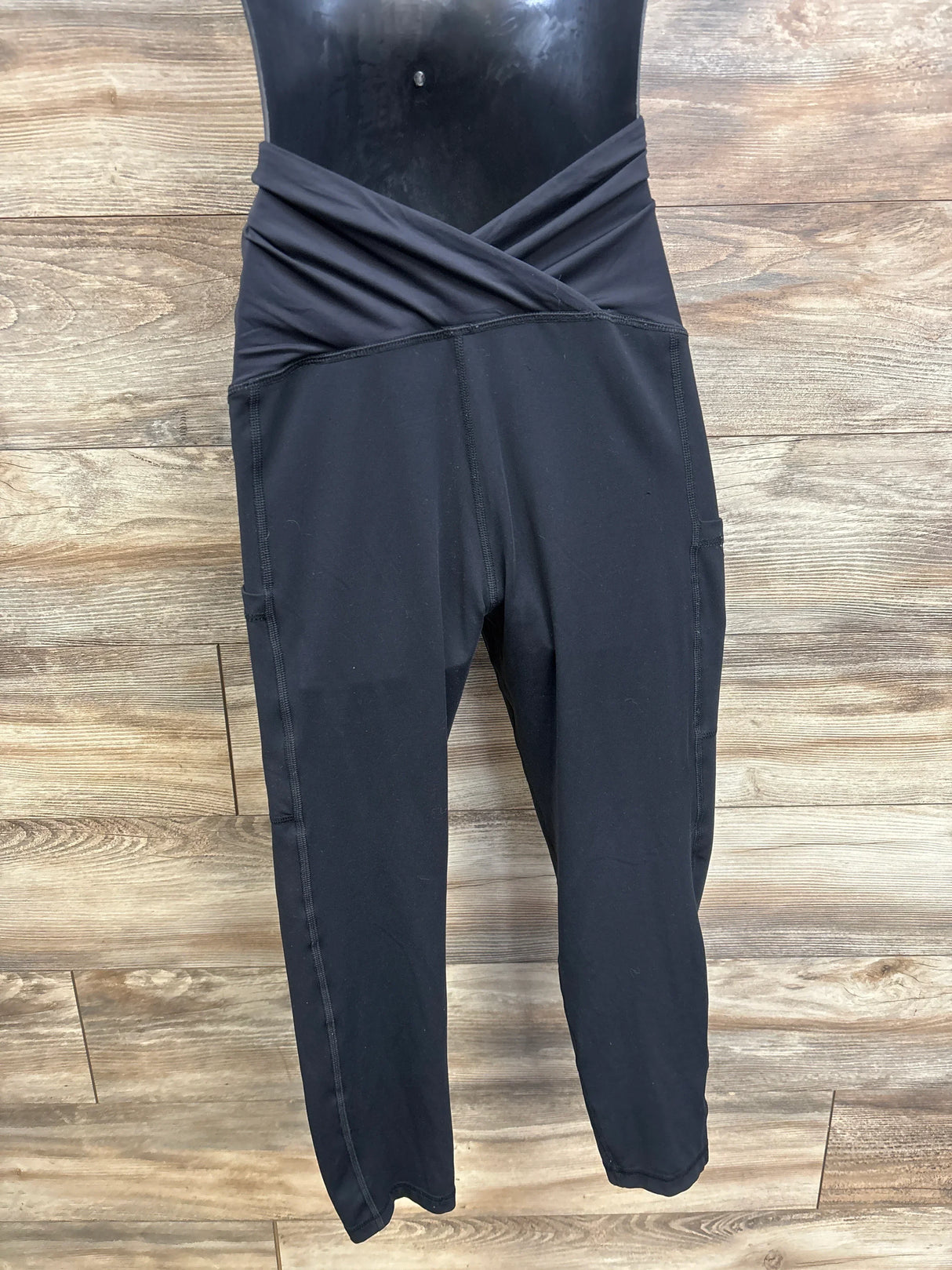 Front view of Isabel Maternity black capri leggings on mannequin showcasing crossover waistband and side pockets with wood plank background
