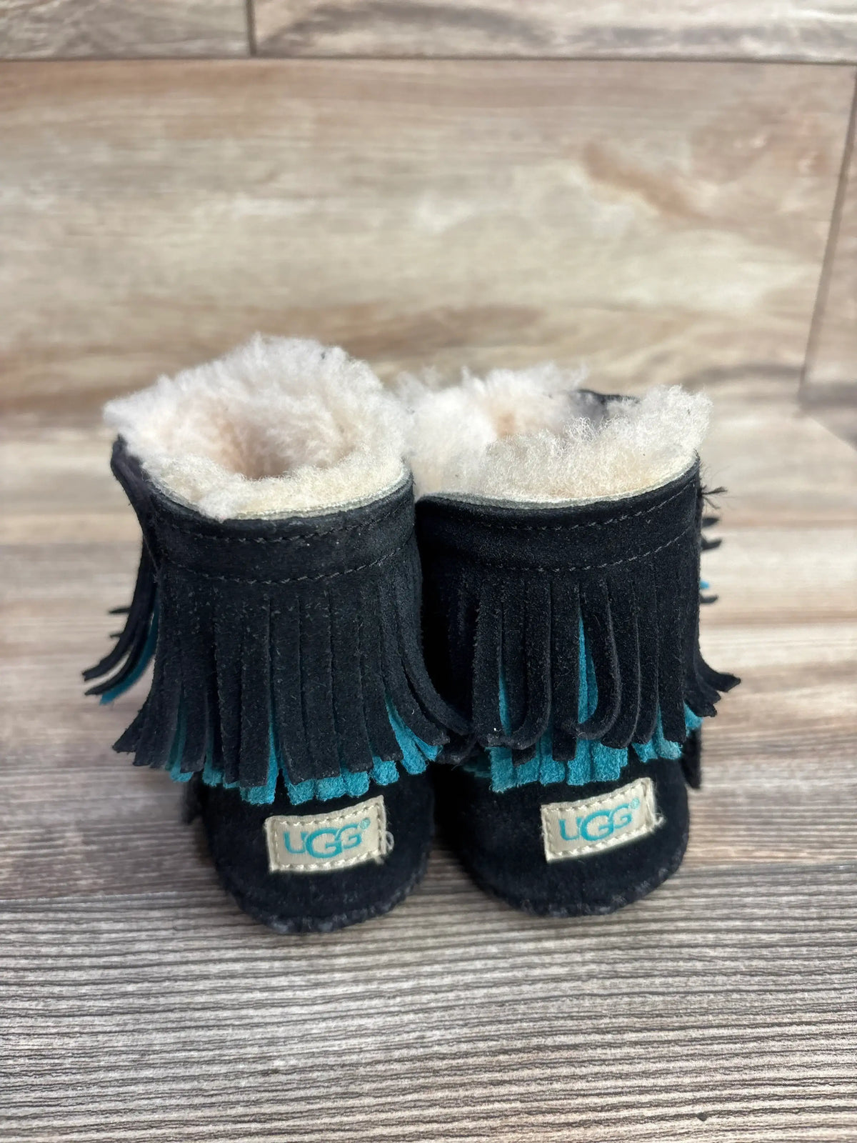 Black UGG baby boots with teal and black fringe, soft wool lining, on wooden surface