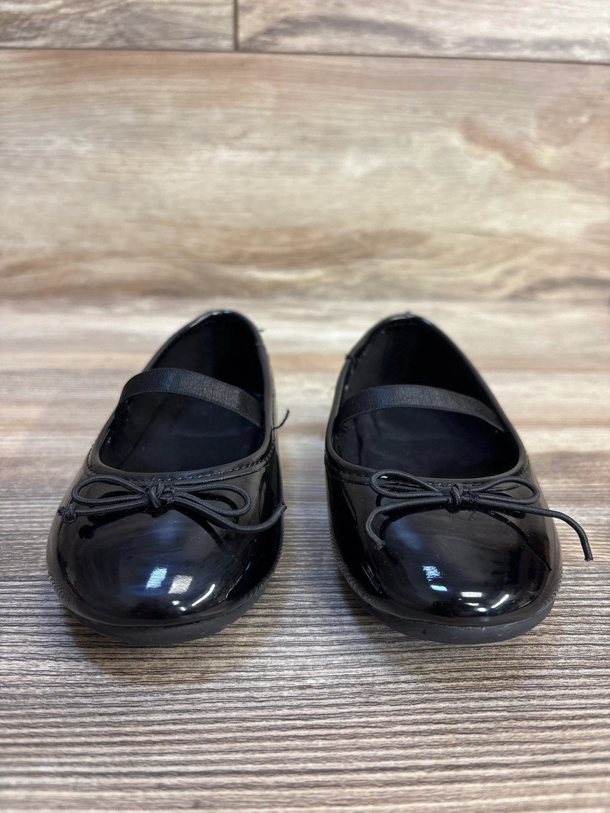Black patent leather toddler dress shoes with bows and elastic straps on wooden floor