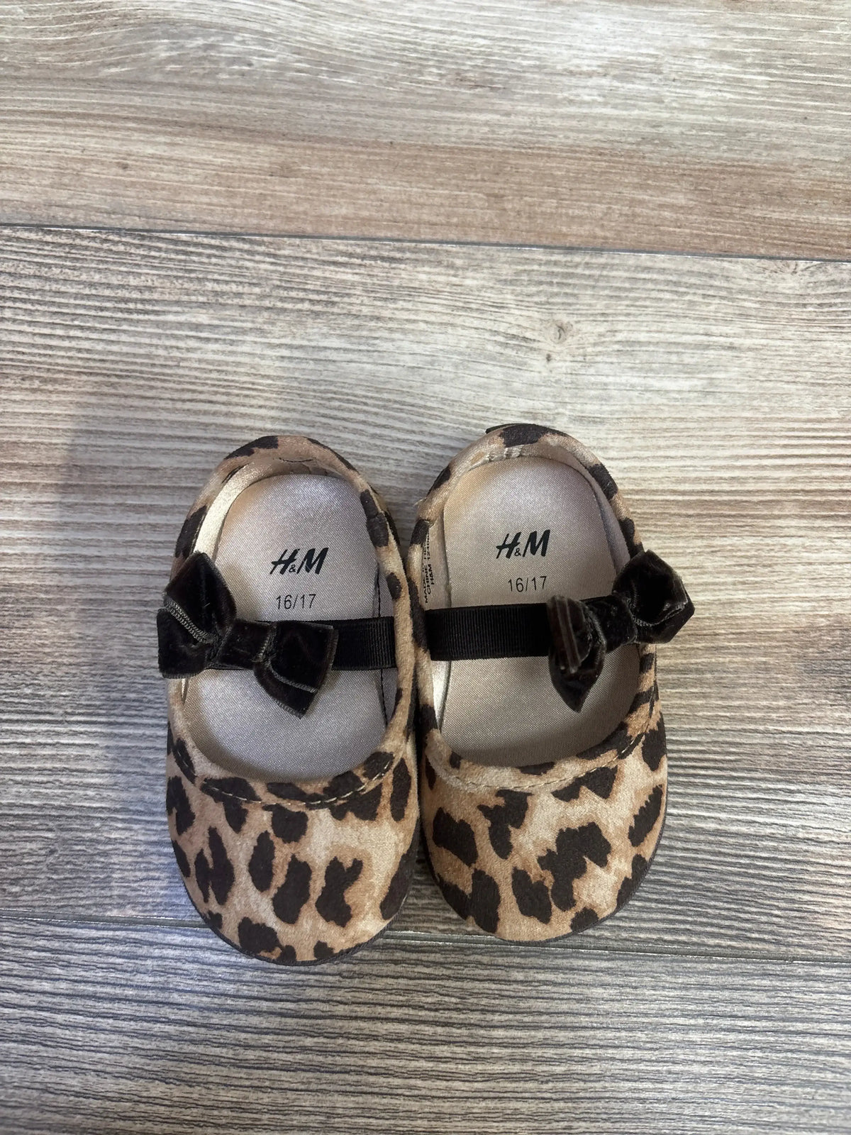Top-down view of H&M infant leopard-print ballet flats with velvet bows on light wood floor, insoles marked 16/17