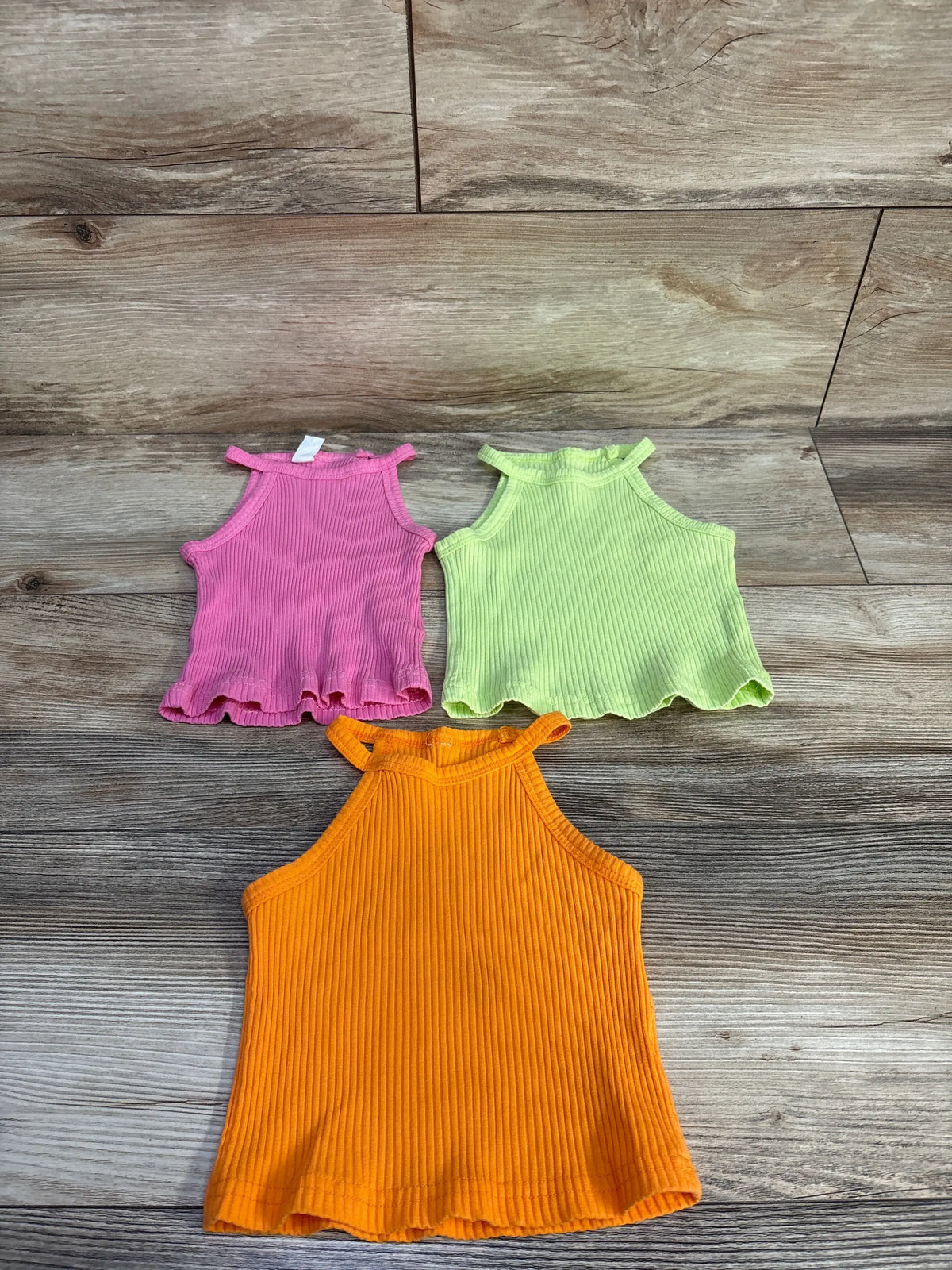 Top view of 3pk ribbed tank tops in pink, lime green, and orange showcasing ruffled hems on a wood-grain background