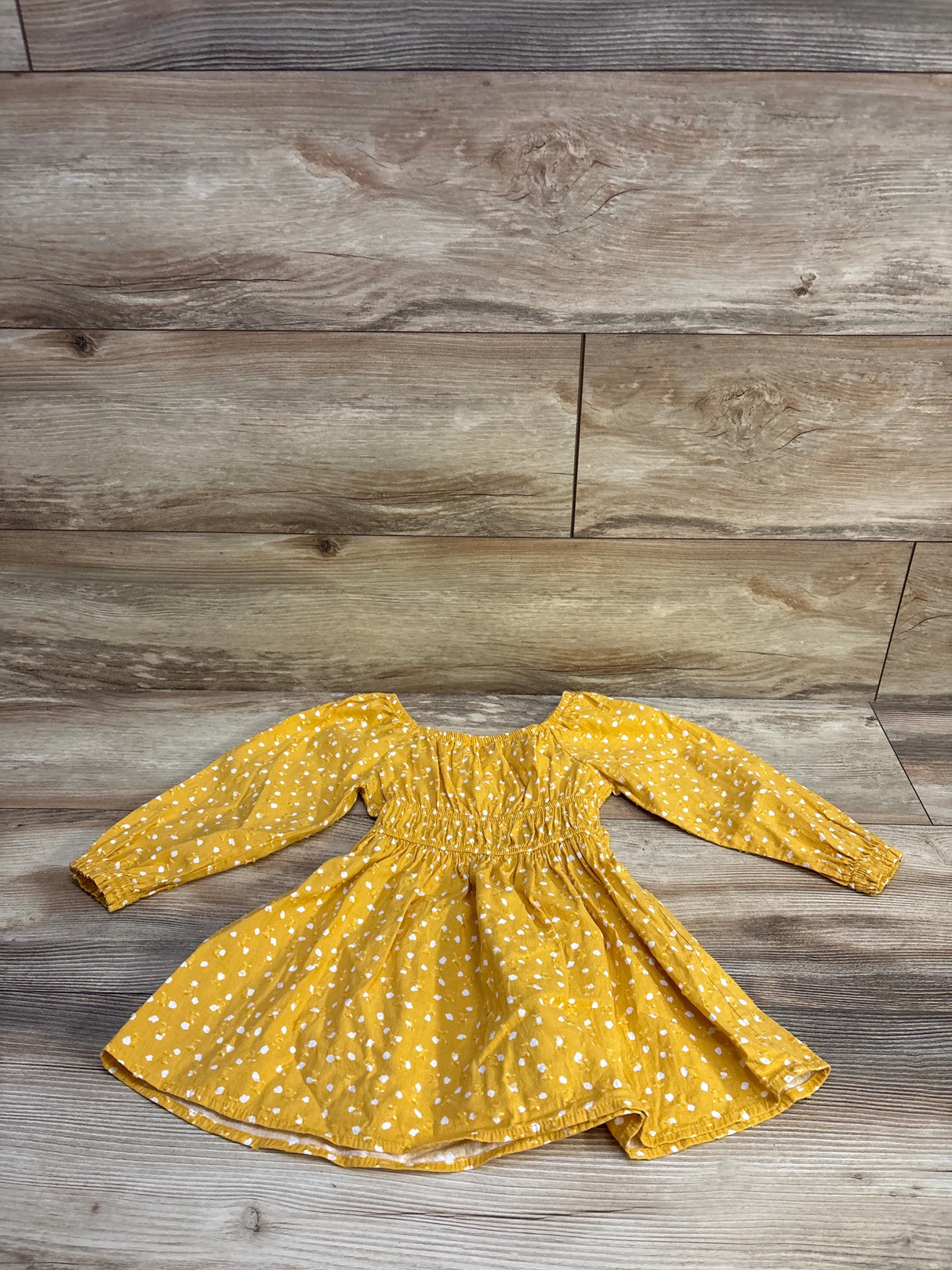 Flat lay of long-sleeve toddler dress showcasing smocked waist and white Swiss dot print with wood plank background