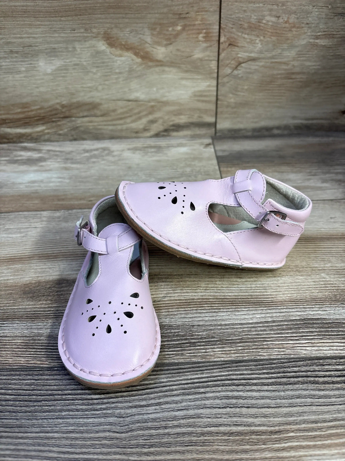 Front and side view of pink leather Cole Haan toddler Mary Jane shoes with floral cutouts, ankle buckle strap, and tan stitched sole on wood background