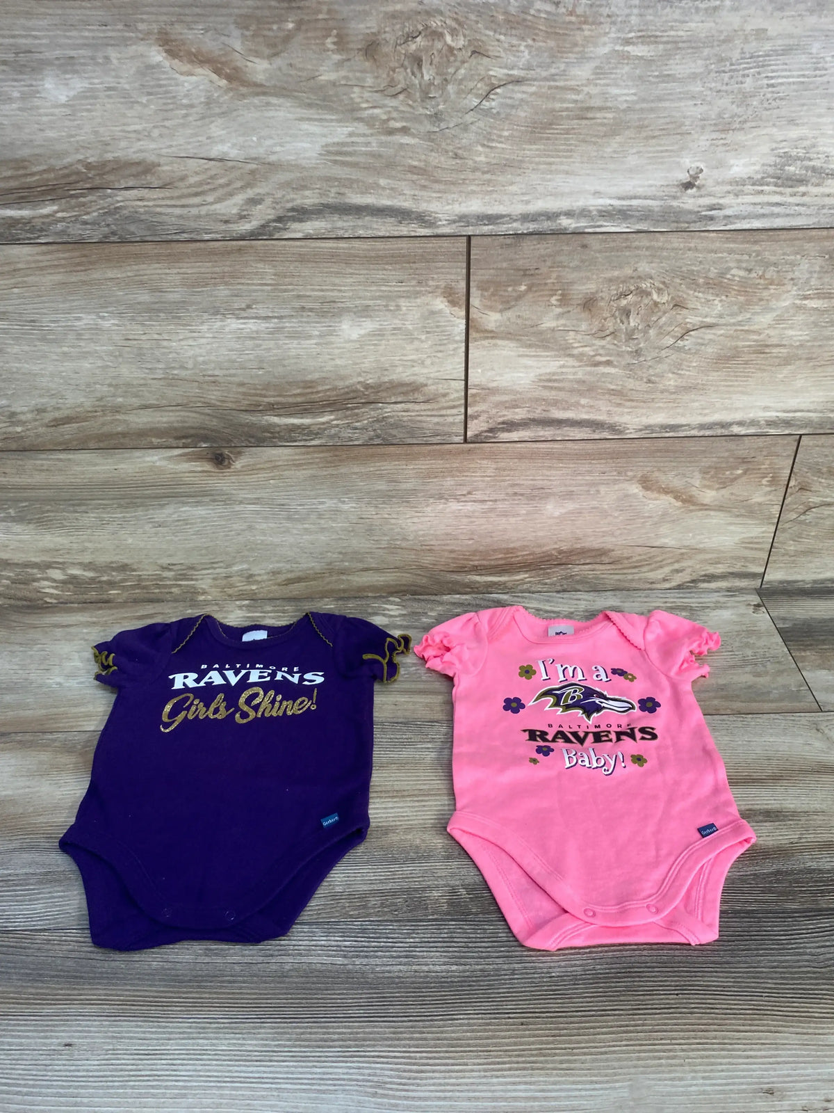 Front view of two Baltimore Ravens bodysuits, purple and pink, with team graphics and snap closures on a wood-grain floor