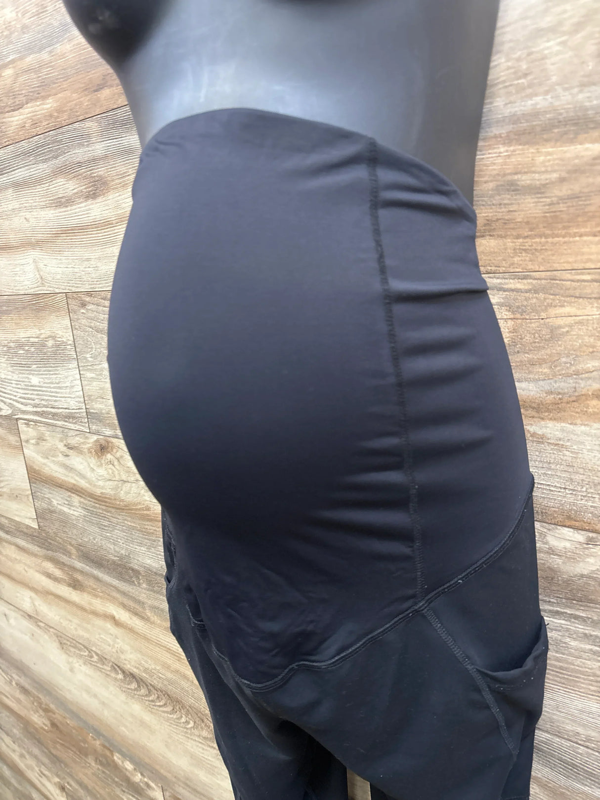 Side view of Isabel maternity capri leggings in black showing high over-bump belly panel, capri length and thigh pocket on wood background