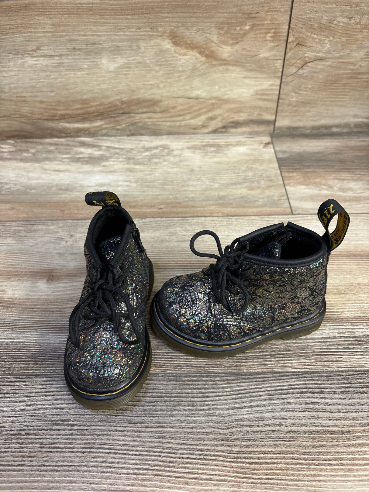 Front view of toddler Dr. Martens metallic lace-up boots showing glitter crackle upper and yellow welt stitching on wood