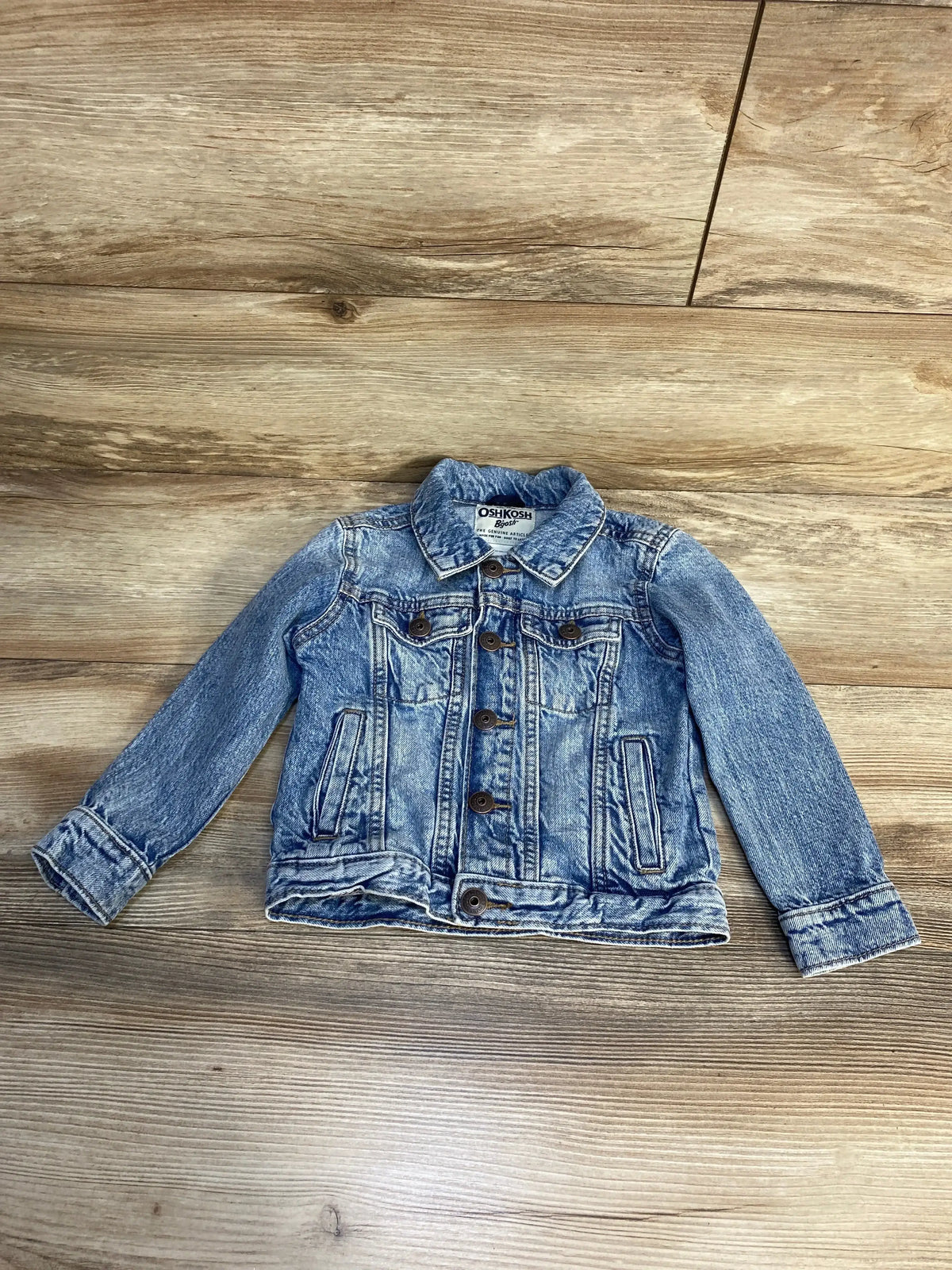 Front view of OshKosh light blue toddler denim jacket (2T) showing button-front and chest pockets on wood floor
