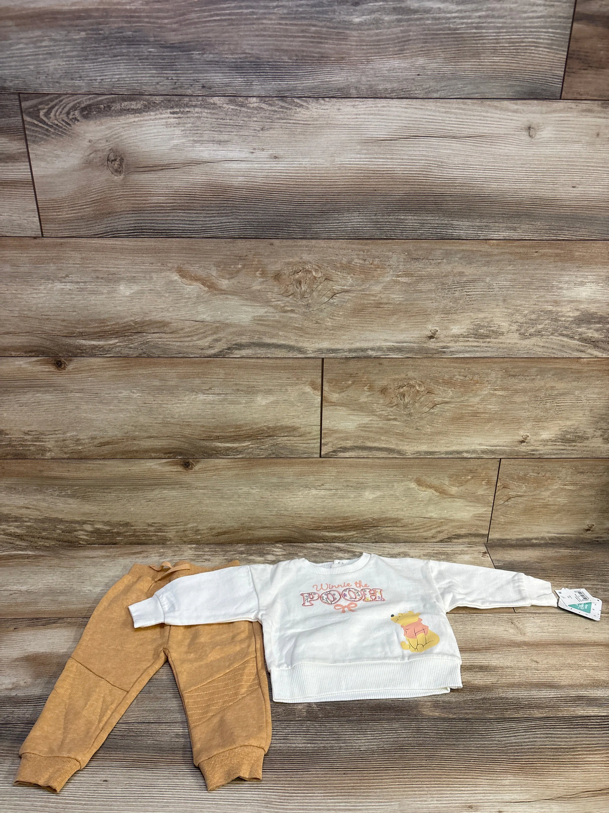 Flat lay of Disney Baby Winnie the Pooh white sweatshirt with Winnie the POOH text and Pooh graphic paired with tan joggers, 6-9M on light wood floor