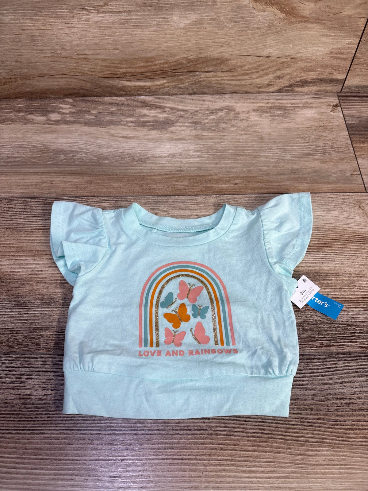 Top-down view of teal 3m baby shirt showing ruffle sleeves and rainbow-and-butterflies graphic reading LOVE AND RAINBOWS