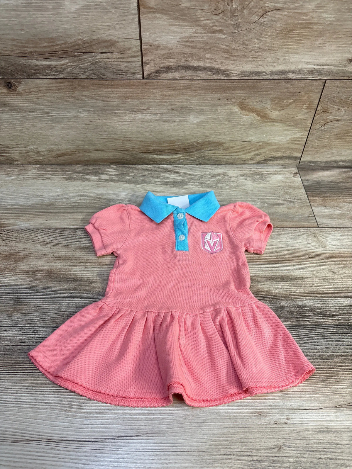 Front view of coral Vegas Golden Knights A line polo dress with blue collar, chest logo, and short sleeves on wood floor