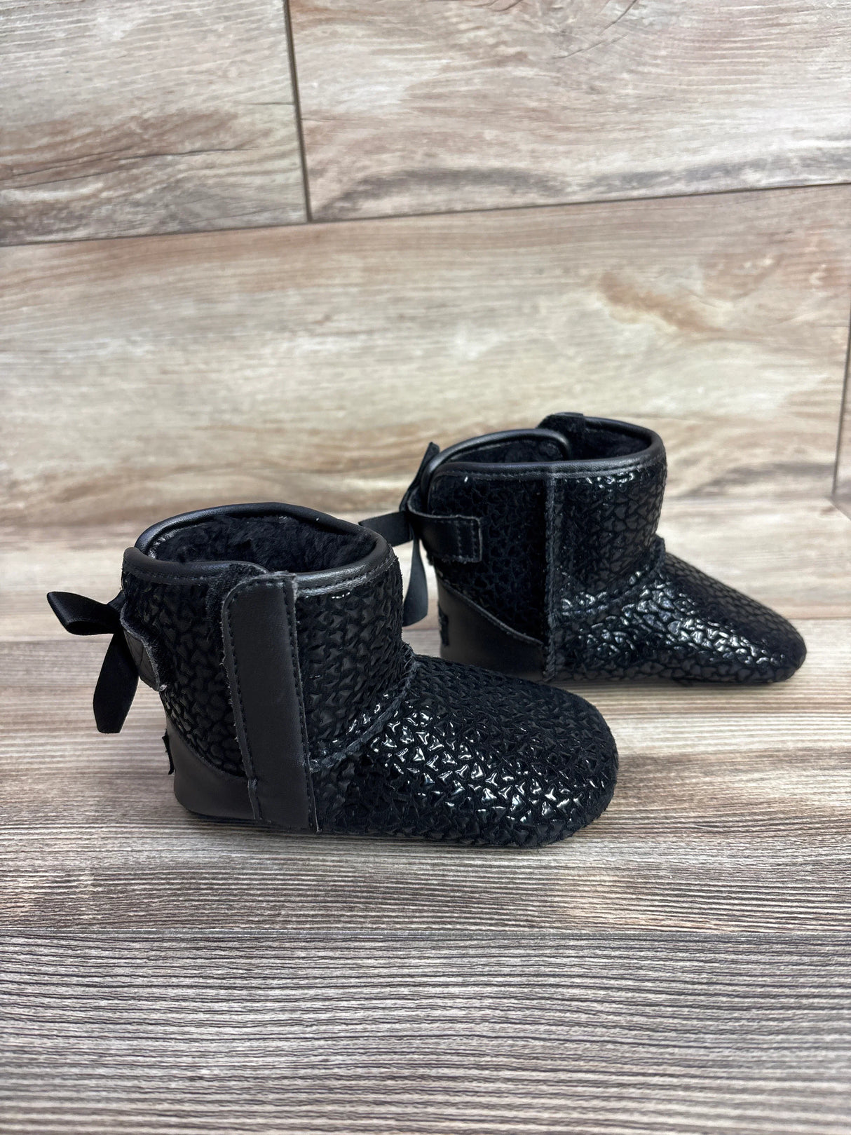 Side view of Ugg Jesse Bow II Gel Hearts baby boots in black showing textured upper, back ribbon bows, and plush lining on wood floor