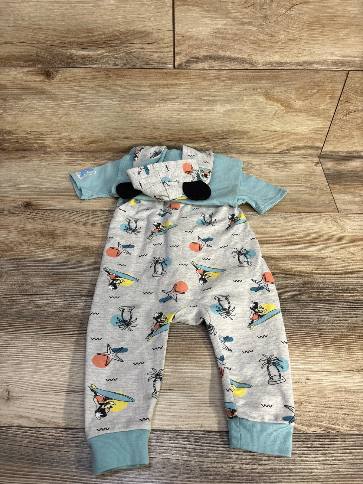 Flat lay of Disney Baby Mickey Mouse hooded zip-up jumpsuit showcasing grey patterned fabric and teal cuffs on a wood floor