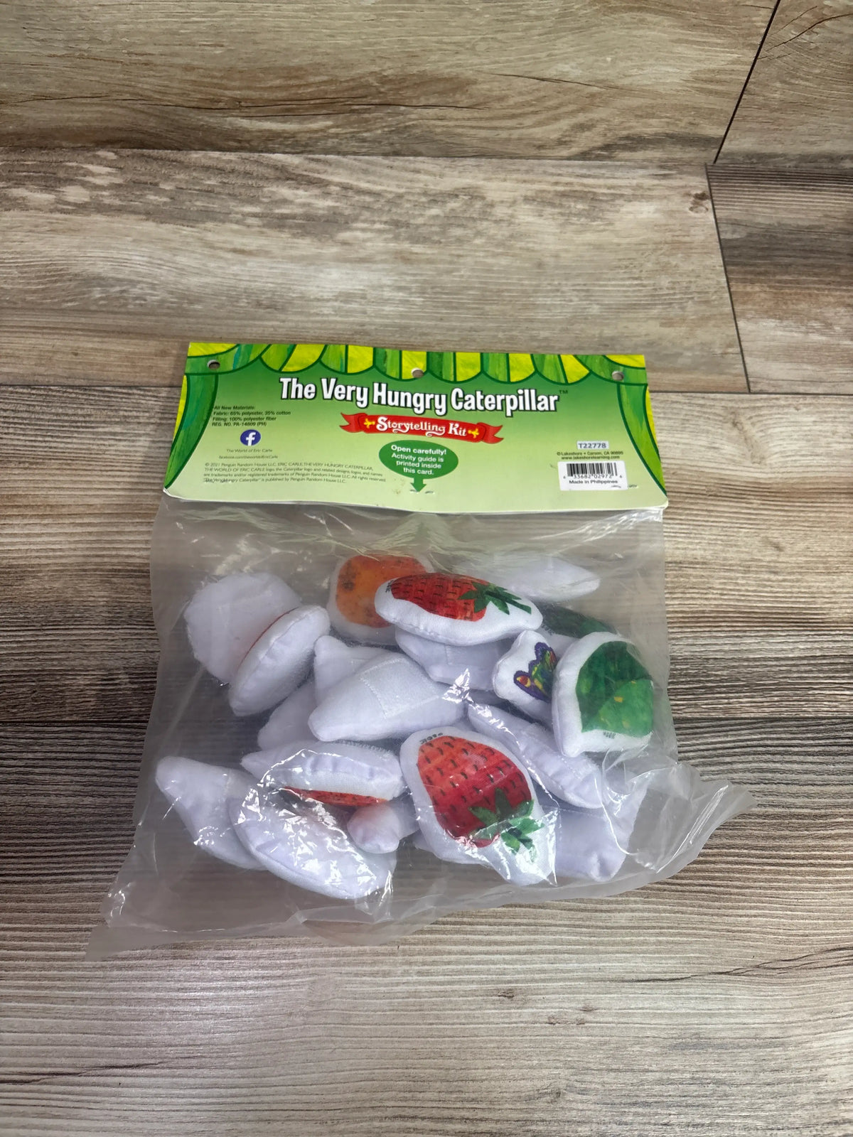 Top-down view of The Very Hungry Caterpillar Storytelling Kit showcasing fruit-themed storytelling pieces in a clear bag on a wood surface