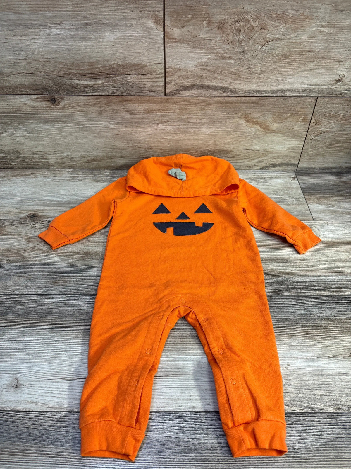 Flat lay of orange pumpkin hooded romper showing jack-o’-lantern face, hood with green stem, and snap leg closures on wood floor