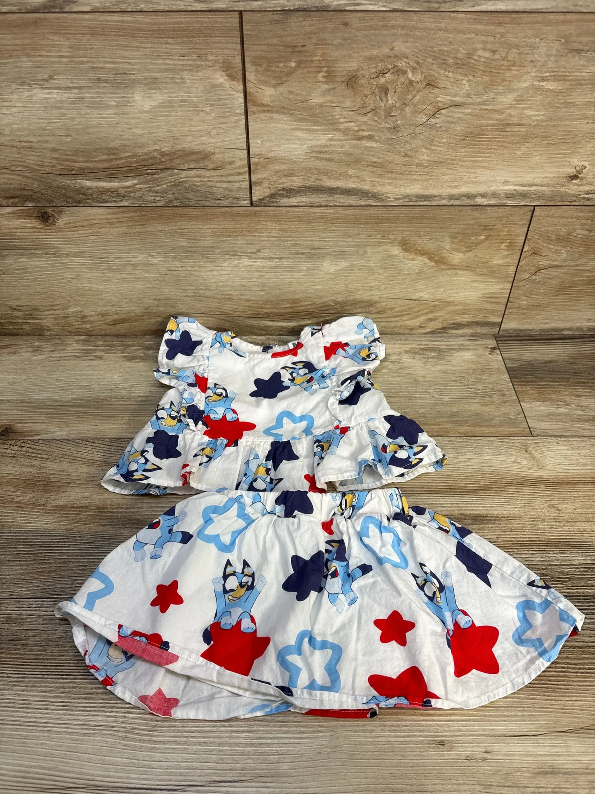 Top-down of girls Bluey peplum top and skort two-piece set, white, showing Bluey print and red and blue stars on wood floor