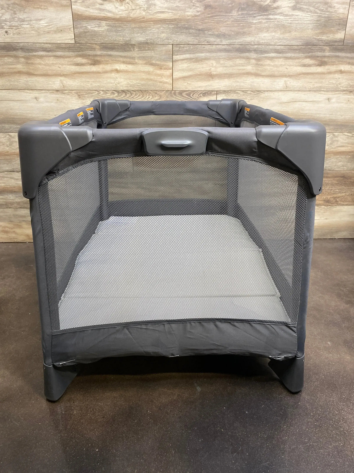 Front view of 4moms Breeze Go playard showing mesh sides and mattress with wood-plank wall and dark floor background