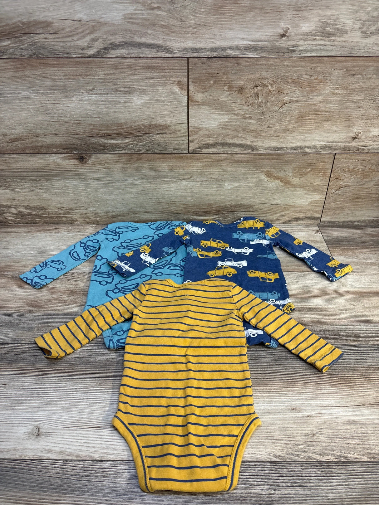 Flat lay of three Carter's long-sleeve bodysuits, 9M, mustard striped, navy car print, and light blue car outlines on wood