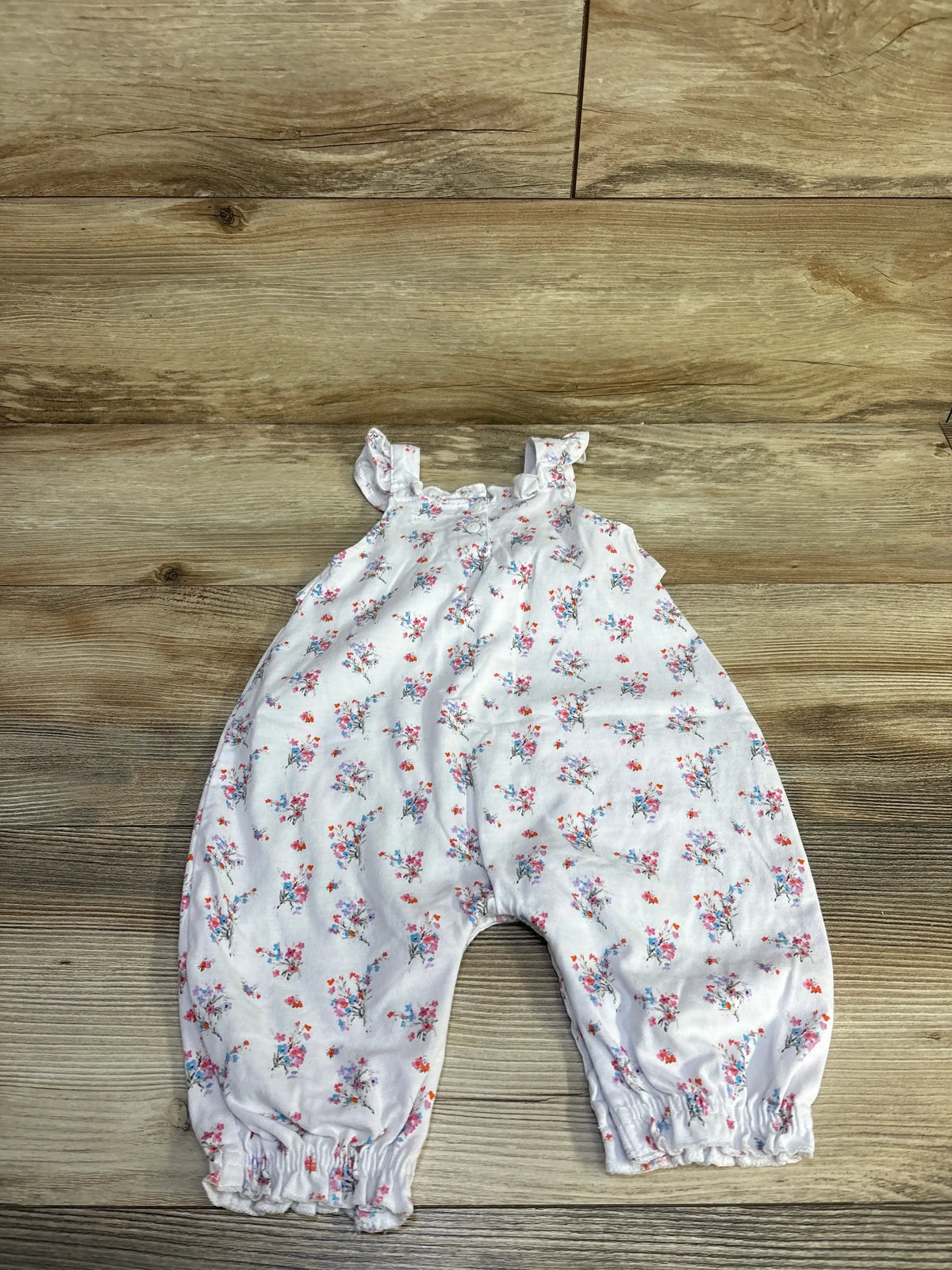 Flat-lay front of Nannette baby jumpsuit, white floral print with ruffle straps and elastic ankles on wood background