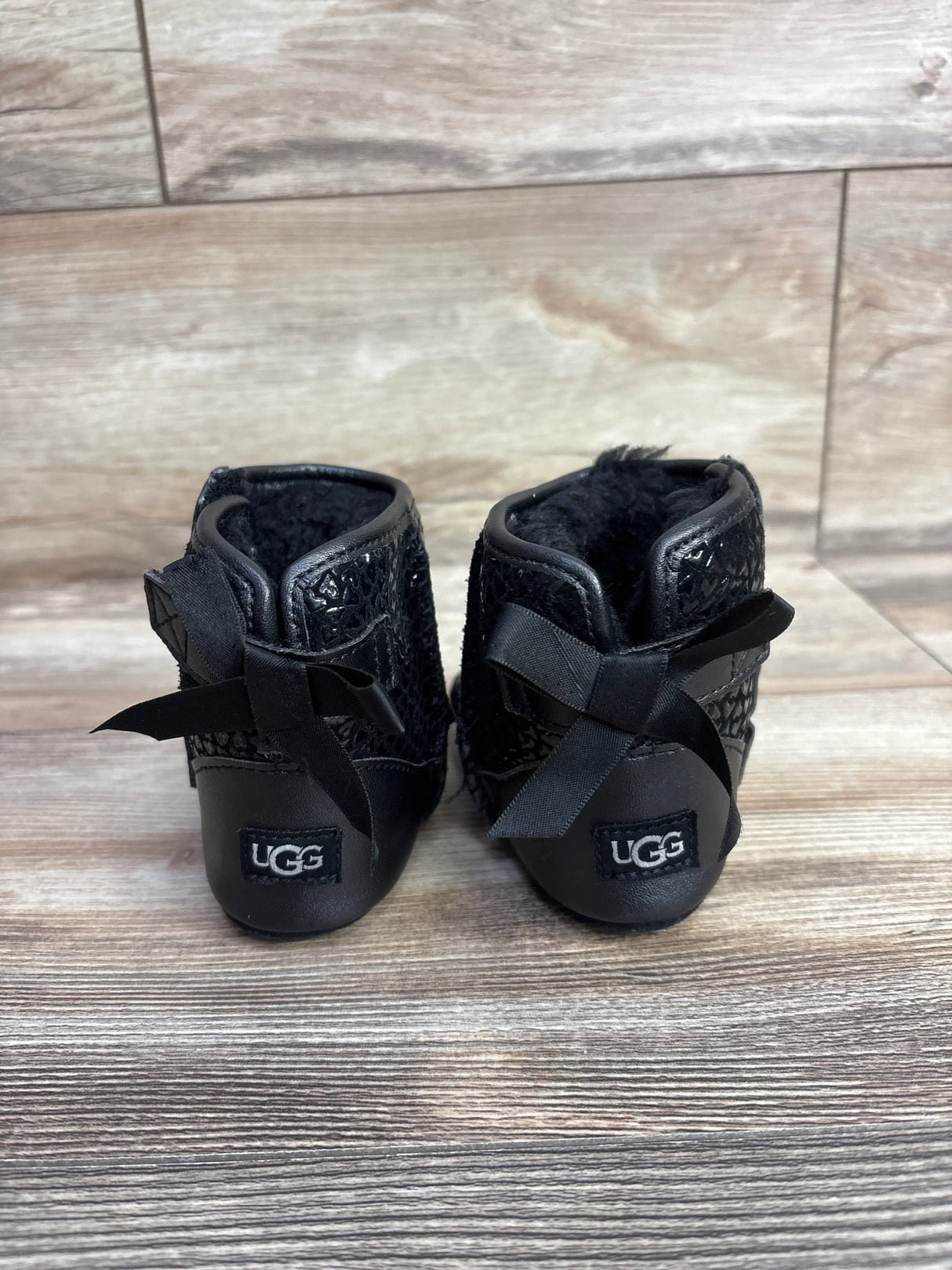 Pair of black toddler UGG boots with bows on the back, displayed on wood background